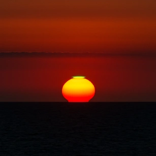 A vivid sunset over the ocean with a rare green flash visible at the top edge of the setting sun, surrounded by deep red and orange sky and dark sea below.
