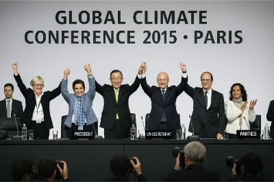 Diverse delegates on a conference dais in Paris, 2015, raise joined hands before a backdrop reading ‘Global Climate Conference 2015 • Paris’; nameplates such as ‘President’ and ‘UN Secretariat’ sit on the table.
